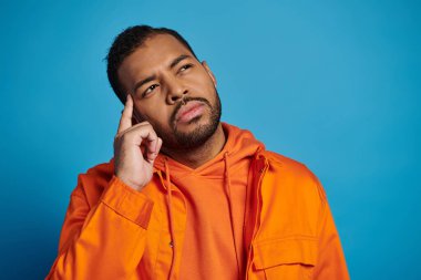 20 'li yaşlarındaki düşünceli Afrikalı Amerikalı adam mavi arka planda parmaklarıyla tapınağa dokunuyor.