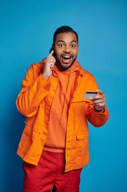 Neşeli Afrikalı Amerikalı adam kredi kartıyla akıllı telefondan arıyor ve kameraya bakarken şaşırıyor.