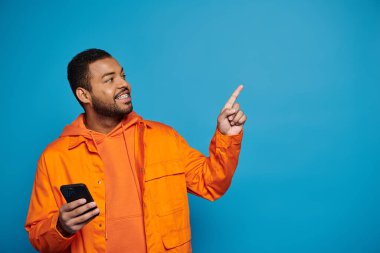 Turuncu elbiseli yakışıklı Afro-Amerikan genç adam akıllı telefonuyla parmağını kaldırıyor.