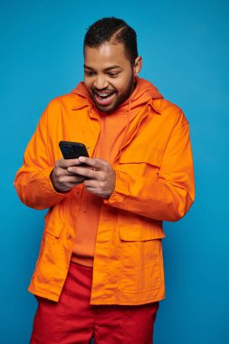 Turuncu kıyafetli Afrikalı Amerikalı oyuncu mavi arka planda akıllı telefondan oyunu kazanıyor.