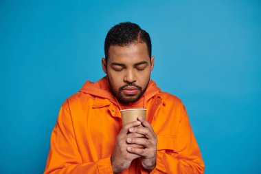 Turuncu kıyafetli donmuş Afro-Amerikan adam elinde mavi arka planda kağıt bardakla