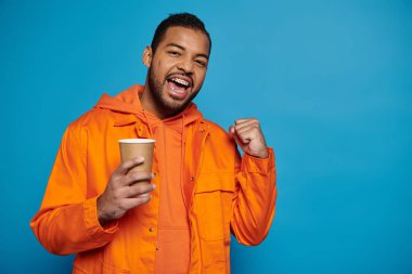 20 'li yaşlarda, mutlu Afro-Amerikan bir adam elinde kağıt bardakla mavi arka plana yumruk atıyor.