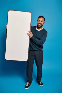 Çekici Afrikalı Amerikalı genç adam mavi arka planda elinde akıllı telefon modeli tutuyor.