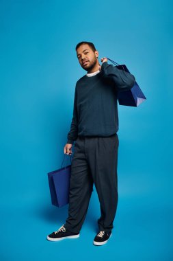Koyu mavi kıyafetli Afro-Amerikan adam. Elinde parlak bir arka planda alışveriş torbalarıyla poz veriyor.
