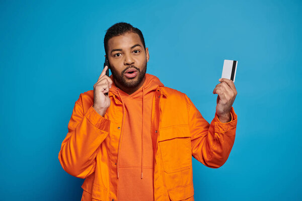 surprised african american young guy calling in smartphone with credit card on blue background