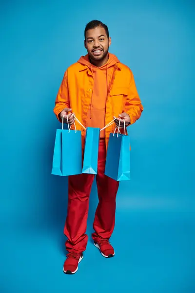 Wesoły Afroamerykanin Mężczyzna Pomarańczowym Stroju Trzyma Torby Zakupy Niebieskim Tle — Zdjęcie stockowe