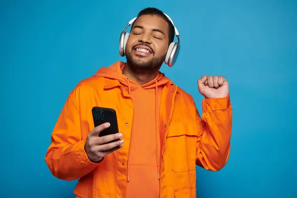 stock image cheerful african american man in orange outfit and headphones dancing with smartphone