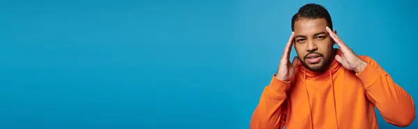 stock image banner of african american man in orange outfit touching to temple with hands on blue background