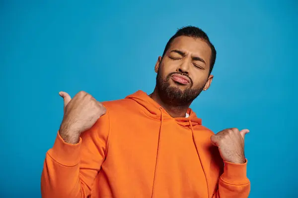 stock image african american man with closed eyes outfit pointing away with thumbs on blue background