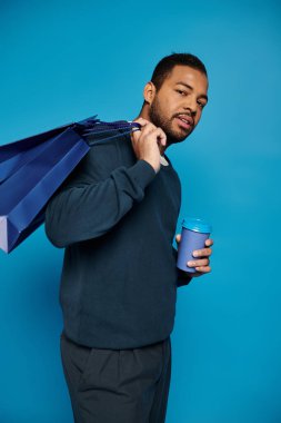Koyu mavi kıyafetli yakışıklı Afro-Amerikan bir adam elinde çantasıyla omzunun üzerinde alışveriş torbaları tutuyor.
