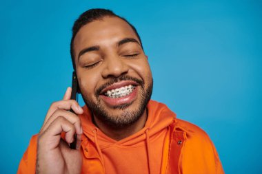 Yakışıklı Afrikalı Amerikalı, mavi arka planda kapalı gözleri olan akıllı telefondan arıyor.