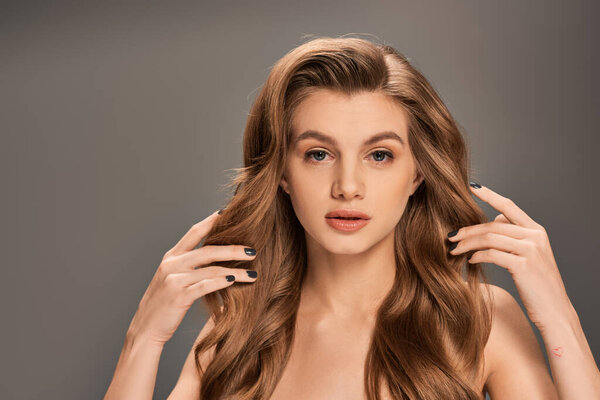 Молодая красивая женщина с длинными волнистыми каштановыми волосами держит руки на голове и волосах.
