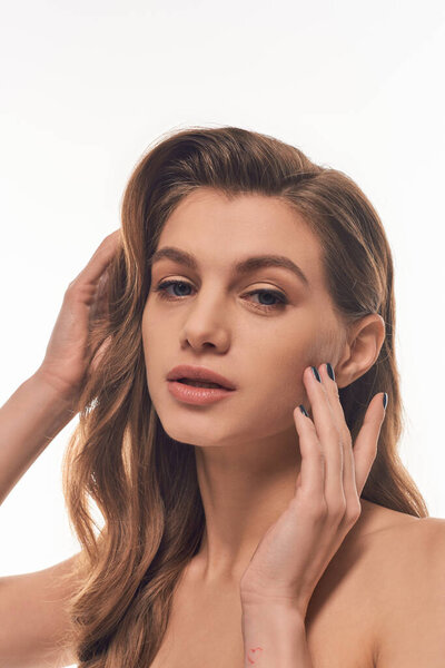 A young woman with long wavy hair gently touches her face.