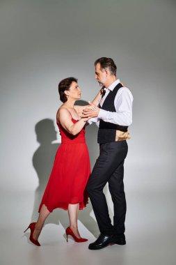 Kırmızı elbiseli ve takım elbiseli, gri arka planda dans eden olgun ve çekici bir çiftin tam boy görüntüsü.