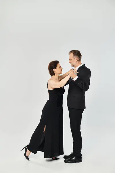 A woman in black dress and man in dark suit embracing on grey background, in tango pose