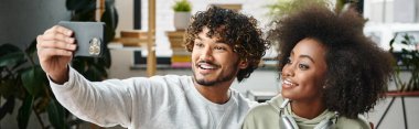 A man and woman of different cultural backgrounds smile while taking a selfie with a cell phone in a modern coworking space. clipart