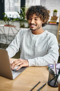 Çeşitli bir gruptan bir adam bir masada oturuyor, modern bir çalışma alanında dizüstü bilgisayarının ekranına gömülü..