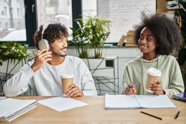 A man and woman of different cultures sitting at a table, sipping coffee and engaging in conversation in a modern coworking space. clipart