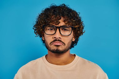 An Indian student with curly hair and glasses poses confidently against a blue backdrop in a studio setting. clipart