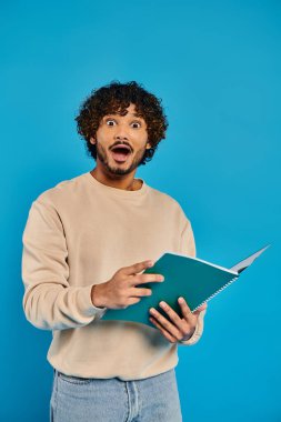An Indian student with curly hair holding a book, standing casually against a blue backdrop in a studio. clipart