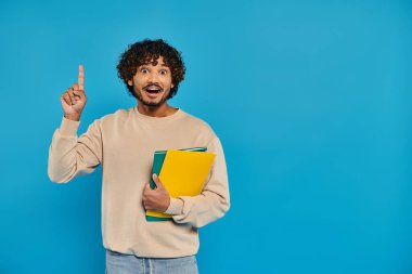 Sıradan giyinmiş Hintli bir öğrenci elinde bir klasörle stüdyoda mavi bir arkaplanı işaret ediyor..