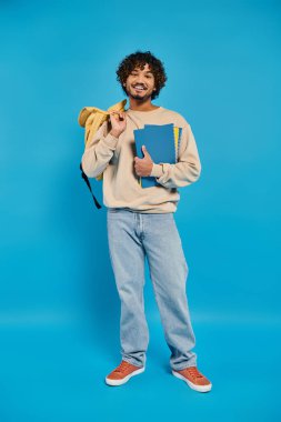 Sırt çantası ve kitap tutan genç bir Hintli öğrenci mavi bir arkaplana karşı günlük kıyafet içinde..