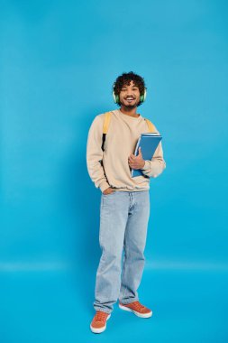 An Indian student standing in casual attire, holding a book in his hands against a blue backdrop in a studio. clipart