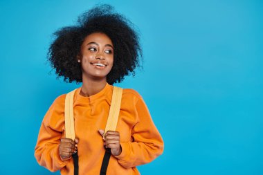 Afro-Amerikalı bir üniversiteli kız, omuzunda bir pantolon askısı ve mavi bir arka plan ile rahatça ayakta duruyor..