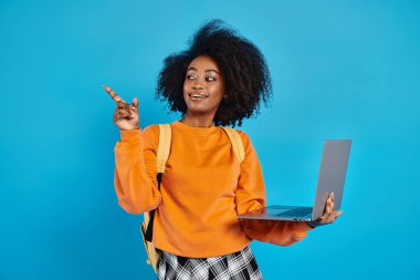 Sıradan giyinmiş, elinde dizüstü bilgisayarla, mavi arka planlı bir stüdyonun yan yüzünü gösteren Afrikalı bir üniversiteli kız..