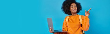 Afro-Amerikalı bir üniversiteli kız, elinde dizüstü bilgisayarla mavi bir arkaplanda dikiliyor..