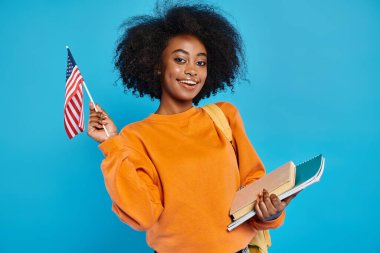 Afrikalı bir Amerikalı üniversiteli kız gururla bir stüdyo ortamında kitap ve Amerikan bayrağı tutuyor..