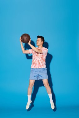 Moda giyinmiş şık bir genç adam enerjik bir şekilde sağ elinde mavi bir fona karşı bir basketbol topu tutuyor..