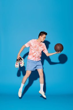 Modayı takip eden genç bir adam canlı mavi bir arka planda basketbol topuyla poz veriyor..