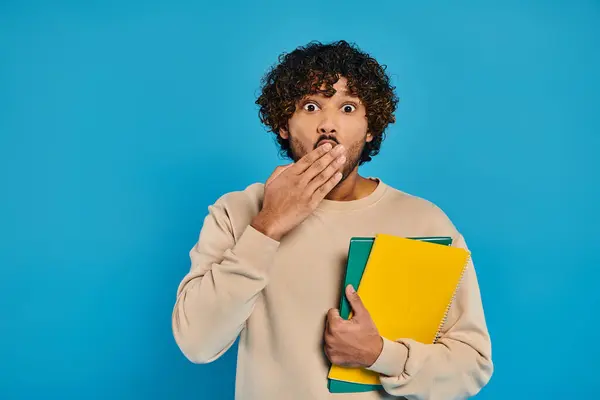 stock image A man of Indian descent stands in casual attire against a blue backdrop, holding a folder and displaying a surprised expression.