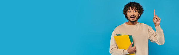 An Indian student stands in casual attire, holding a book and pointing upwards against a blue backdrop.