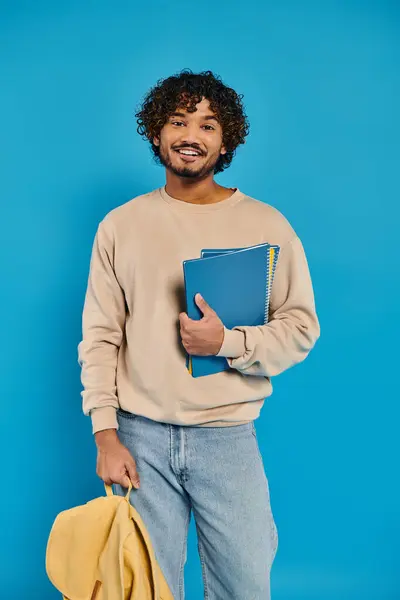 一个留着卷发的印度年轻人拿着一本书 站在工作室的蓝色背景下 — 图库照片