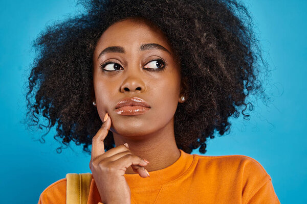 A stylish African American college girl gazes upwards with her stunning afro hair against a blue backdrop in a studio.