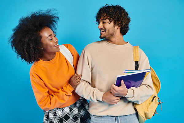 An interracial man and woman stand side by side in casual attire against a blue backdrop in a studio setting.