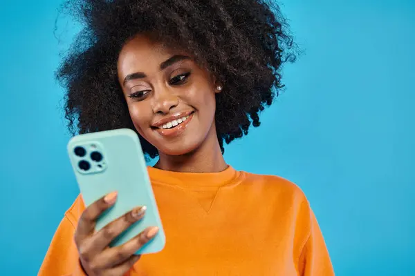A black student in casual attire smiling while holding a cell phone on a blue backdrop in a studio.