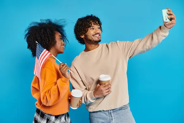 異人種間のカップル スタイリッシュな服装 青い背景 アメリカの国旗に対する携帯電話のセルフィーと一緒に瞬間をキャプチャ — ストック写真