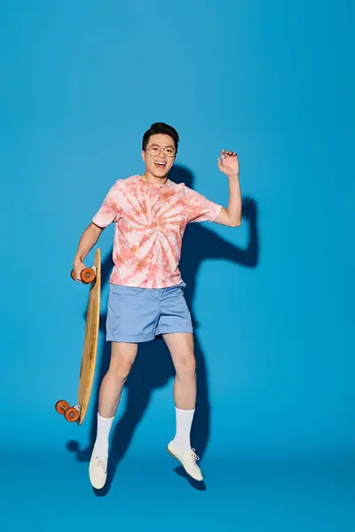 トレンディな服装のスタイリッシュな若者 スケートボードを保持し 青い背景に対してダイナミックなポーズを打つ — ストック写真