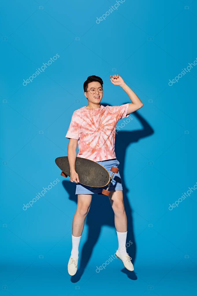A stylish, good-looking young man in trendy attire strikes a pose, holding a skateboard in his right hand on a blue backdrop.