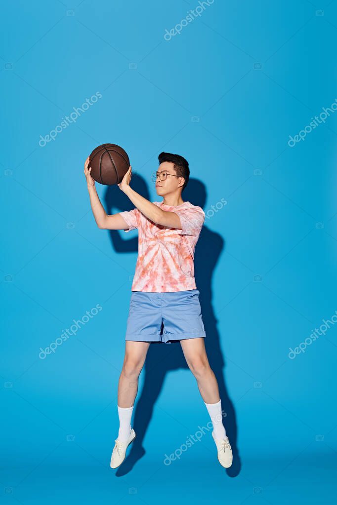 A stylish young man in trendy attire energetically holds a basketball in his right hand against a blue backdrop.
