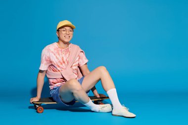 A trendy young man poses stylishly while sitting on a skateboard against a vibrant blue backdrop. clipart