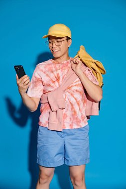 Sırt çantası ve cep telefonu olan şık bir genç adam. Mavi bir arka plana macera ve modern bir bağlantı hissi yayıyor..
