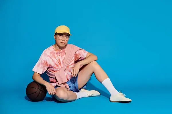 トレンディな服装のスタイリッシュな若者は リラックスとカジュアルな運動の感覚を除いて バスケットボールで地面に座っています — ストック写真