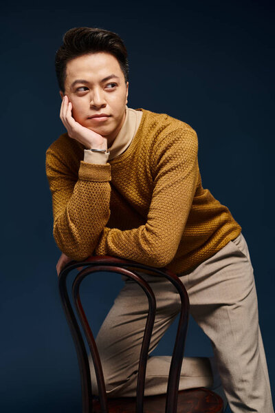Модный молодой человек в элегантной одежде сидит на деревянном стуле в уникальной и уверенной позе.