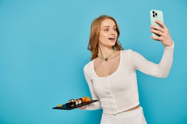 Sarışın kadın elinde lezzetli Asya yemekleri tutarken cep telefonuyla selfie çekiyor ve poz veriyor..