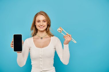 Bir kadın mavi arka planda cep telefonu ve yemek çubuklarını dengeliyor.