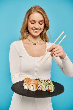 Blonde woman gracefully holds sushi plate and chopsticks, ready to indulge in a delectable Asian meal. clipart